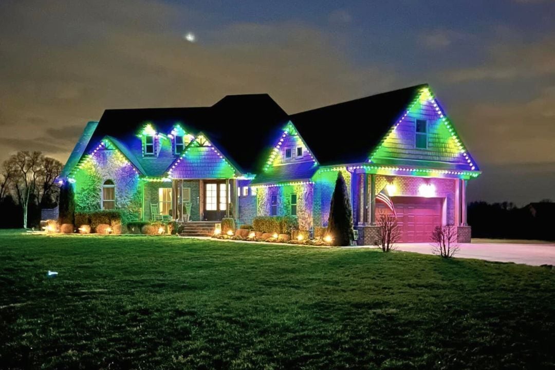 A modern home that has many peaks and features a Trimlight Mardi Gras lighting pattern with a lot of grass and landscape accent lighting.