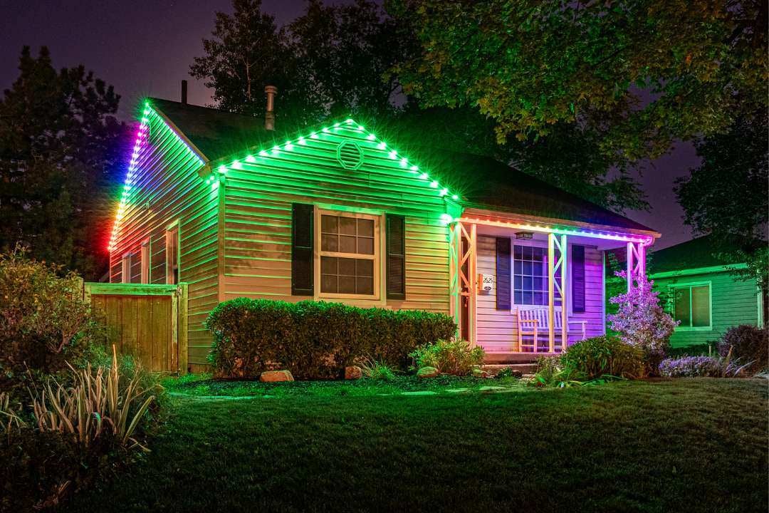 A home that has nice landscape bushes and trees. It is mostly green lighting with a rainbow line of lights in 2 places on the home.