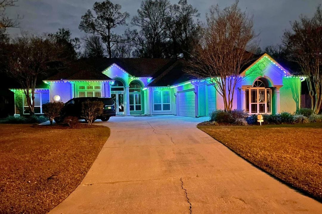 A picture of a home and a car out front with a long driveway. The home has a green and purple lighting pattern for Mardi Gras.